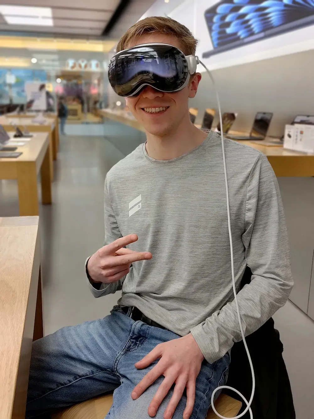 Matt seated in an Apple store wearing Vision Pro. He is smiling and his hand is making a peace sign.