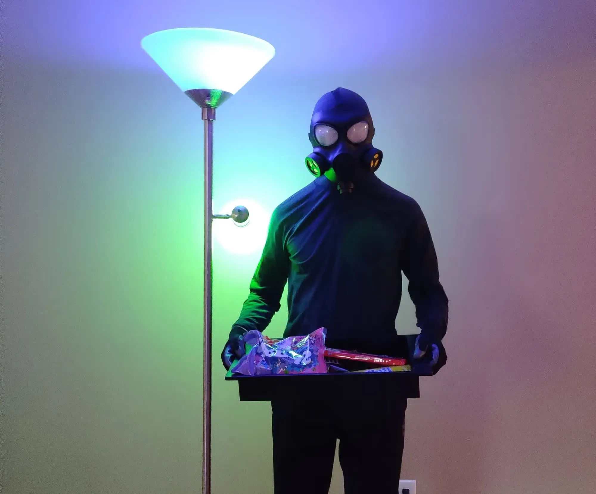 Matt stands inside wearing all black clothes, black gloves, and a black plastic gas mask. He holds a black tray filled with candy. Next to him stands a tall light fixture with his smart bulbs casting blue and green light over him.